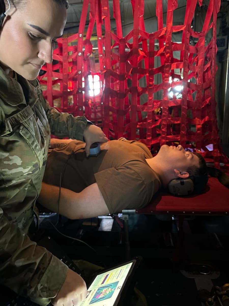 A service member places a handheld sensor on a patient lying in the cargo hold of a C-130 air ambulance, with readings on a tablet