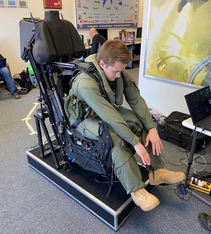 A pilot in a flight suit seated in an ejection seat test rig with a laptop recording beside him
